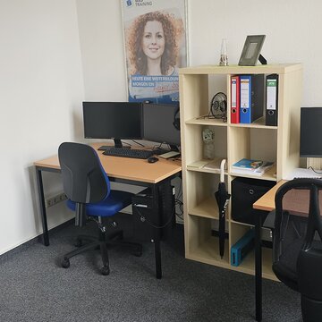2 Arbeitsplätze mit jeweils 2 Bildschirmen, einem Bürostuhl, einer Tastatur und Maus. Darüber hängen 2 Plakate, zwischen den Tischen steht ein Regal mit Ordnern.