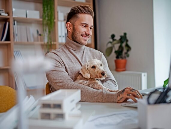 Ein Mann im Homeoffice hat seinen Hund im Arm und arbeitet an seinem Laptop.