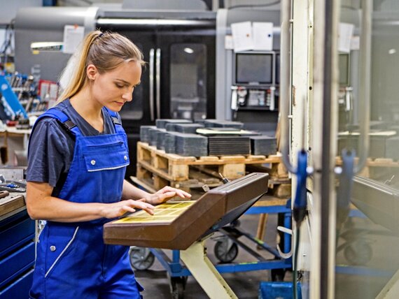 Eine Frau in Arbeitskleidung steht in einer Werkstatt und arbeitet an einer Maschine.