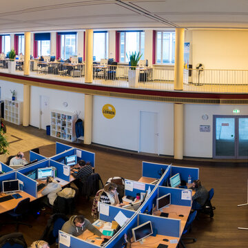 Fisheye-Aufnahme des Arbeitsbereichs und des Aufenthaltwsbereichs. Es befinden sich mehrere, abgetrennte PC-Arbeitsplätze vor Ort. Im Aufenthaltsbereich sieht man mehrere Sofas und Tische an einer Fensterfront.