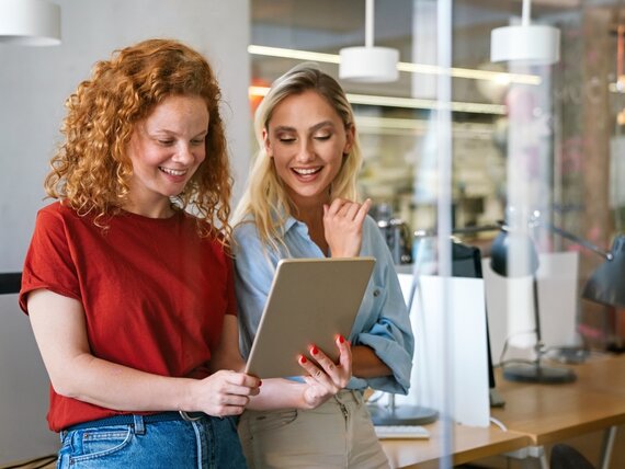Zwei fröhliche Frauen schauen sich etwas auf einem Tablet an.