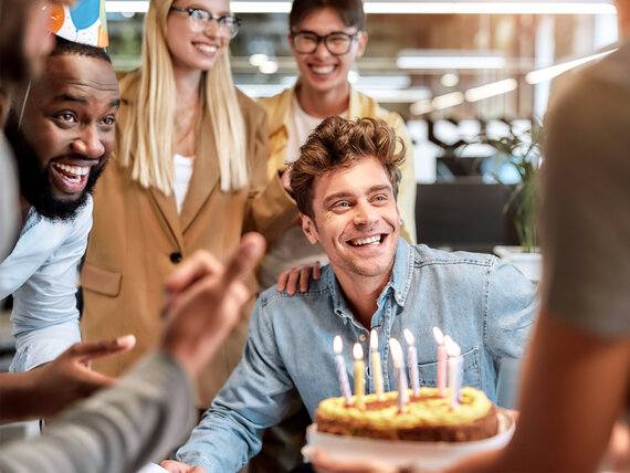 Ein glücklicher junger Mann feiert seinen Geburtstag im Büro mit Kolleg:innen und einer Geburtstagstorte