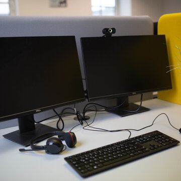 Arbeitsplatz bestehend aus 2 PC-Bildschirmen, Tastatur, Maus und Headset sowie einem blauen Bürostuhl und blauen Trennwänden.