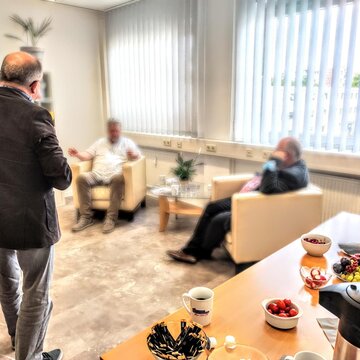 Blick in den hellen Pausenraum in dem ein Tisch mit Kaffee und Snacks steht und Teilnehmende in gemütlichen Sesseln sitzen.