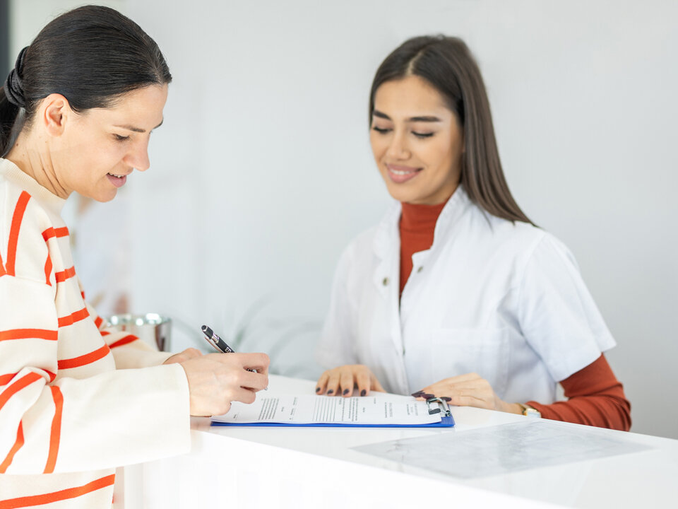 Eine Praxisassistentin hilft einer Patientin am Empfang beim Ausfüllen eines Formulars.