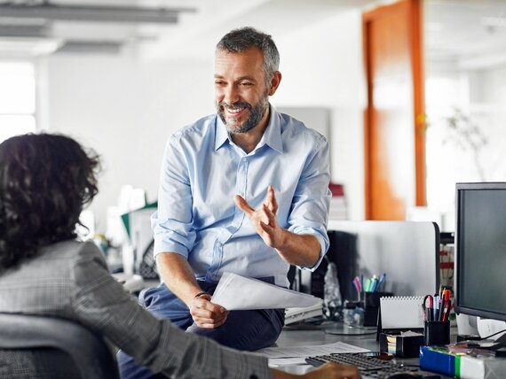 Ein Mann sitzt locker auf einem Schreibtisch im Büro und unterhält sich mit seiner Kollegin.