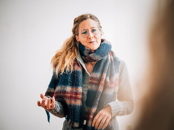 Eine blonde Frau mit Brille und langem Schal hält gerade einen Vortrag