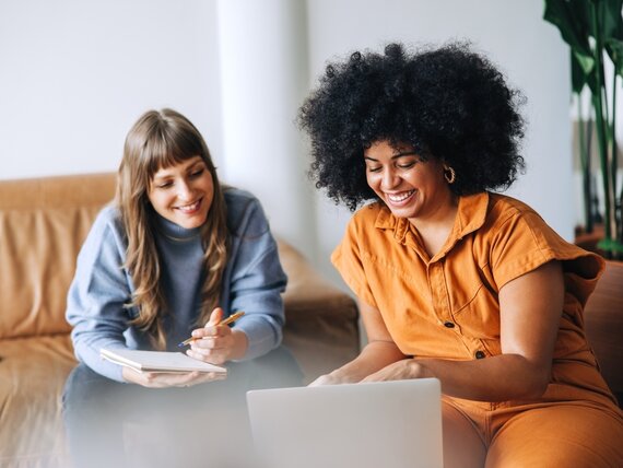 Zwei Frauen sitzen auf einer Couch und besprechen etwas am Laptop. Eine davon schreibt Notizen.