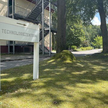 Ein Rasen mit zwei Bäumen vor einem Gebäude. Ein Schild mit der Aufschrift "Technologiezentrum" steht auf dem Rasen.