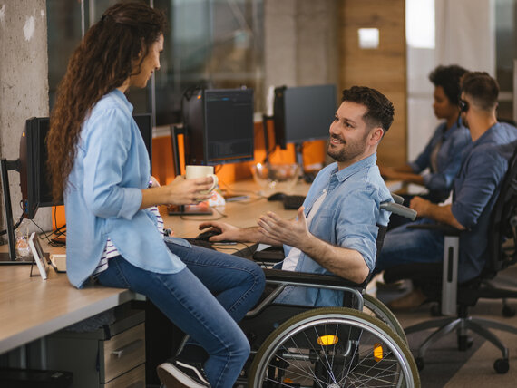 Ein Mann im Rollstuhl, der in einem barrierefreien Büro am Schreibtisch sitzt und sich mit einer Kollegin unterhält.