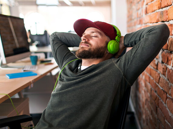 Mann mit Basecap und Kopfhörern sitzt zurückgelehnt im Schreibtischstuhl mit geschlossenen Augen, Arme hinter dem Kopf verschränkt