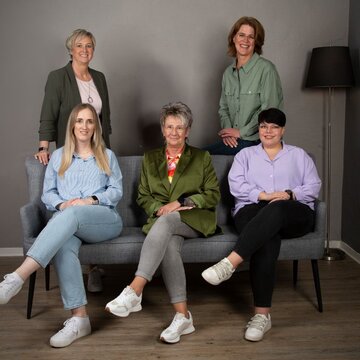 Gruppenbild vom Standortteam. 3 Frauen sitzen auf einem stilvollen, grauen Sofa. Dahinter stehen 2 weitere Frauen. Alle schauen freundlich in die Kamera.