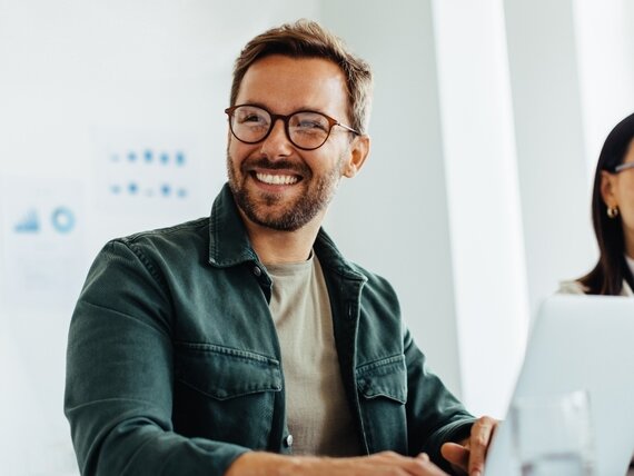Ein fröhlich lachender Mann sitzt neben seiner Kollegin und arbeitet an seinem Laptop.