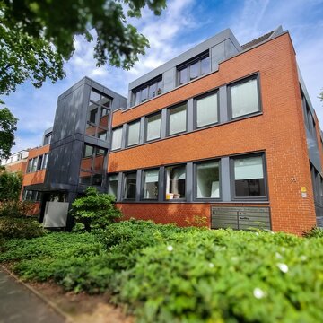 Außenansicht des Schulungsgebäude mit orangenen Klinkern und grauer Fensterumrahmung. Das Gebäude ist umgeben von grünen Pflanzen und Bäumen.