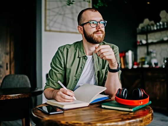 Ein Mann sitzt nachdenklich in einer Küche und schreibt etwas in sein Notizbuch.