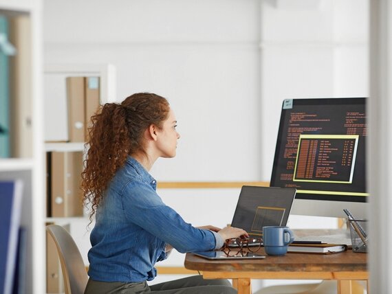 Eine Frau sitzt an einem Schreibtisch mit Laptop und Monitor und arbeitet an Programmiercodes