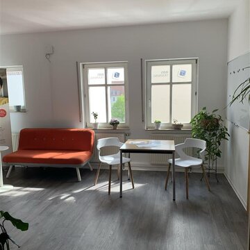 ein Raum mit 3 Fenstern und grauem Boden. Links steht ein rotes Sofa, rechts daneben ein Holztisch mit 2 Stühlen. Hinter dem Stuhl rechts steht eine Zimmerpflanze.