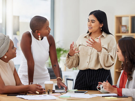 Vier Kolleginnen unterhalten stehen an einem Tisch mit Unterlagen und besprechen etwas.