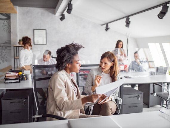 Zwei Frauen sitzen in einem Großraum Büro und besprechen ihre Unterlagen.