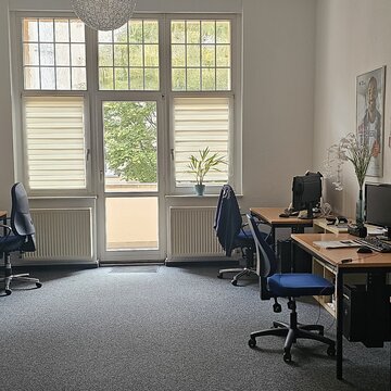 Schulungsraum mit links und rechts mehreren Arbeitsplätzen. Jeweils mit 2 Bildschirmen, einem Bürostuhl, Tastatur, Maus und Headset. Am Ende des Raums mehrere Fenster und eine Tür zum Balkon. An den Wänden mehrere Plakate.
