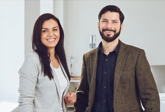 Eine Frau und ein Mann in Businesskleidung stehen nebeneinander und schauen in die Kamera.