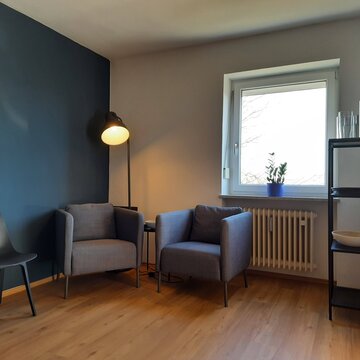 Aufenthaltsraum mit 2 Sesseln vor einer Stehlampe, daneben einem Regal mit Deko und einem weiteren Tisch mit Stuhl