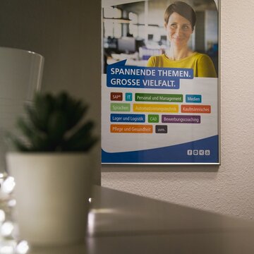 Eingangsbereich mit Poster an der Wand auf dem die Weiterbildungsthemen zu sehen sind. Davor steht ein Regal mit einer kleinen Topfpflanze und einer Lichterkette.