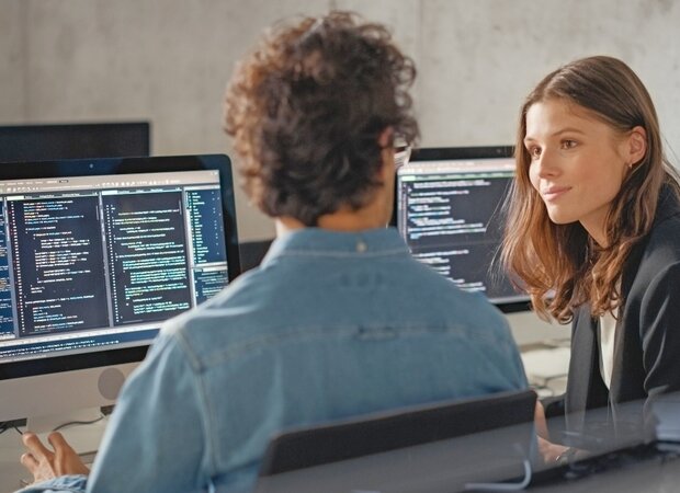 Eine Frau und ein Mann sitzen gemeinsam an ihren Computern auf denen Programmiercodes zu sehen sind.