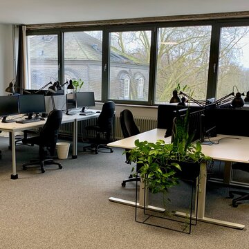 Schulungsraum mit mehreren Arbeitsbereichen, die mit 2 PC-Bildschirmen, Tastatur, Maus und Headset ausgestattet sind. Fensterreihe und einige grüne Zimmerpflanzen.