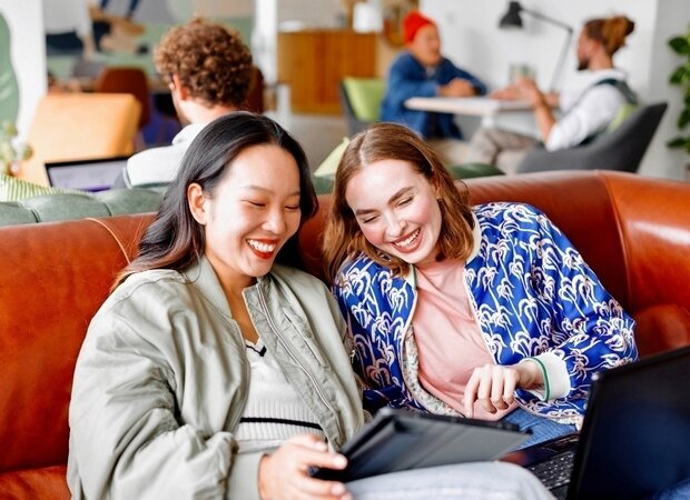 Zwei lachende Frauen sitzen auf einer Ledercouch und schauen sich etwas auf einem Tablet an.