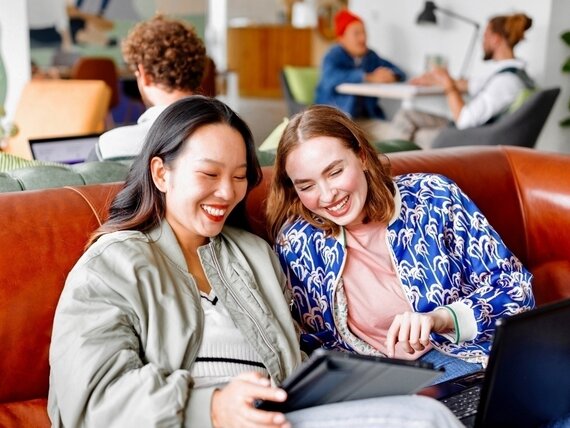 Zwei lachende Frauen sitzen auf einer Ledercouch und schauen sich etwas auf einem Tablet an.