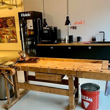Eine moderne, schwarte Küche mit Kaffeemaschine, Wasserkocher und Getränkekühlschrank. Davor steht eine Werkbank aus Holz, die als Tisch dient.