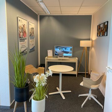 Beratungsbereich mit Tisch und 2 Stühlen. Im Hintergrund ein Regal mit TV, links stehen 2 Pflanzen, an der Wand hängen 2 Plakate.