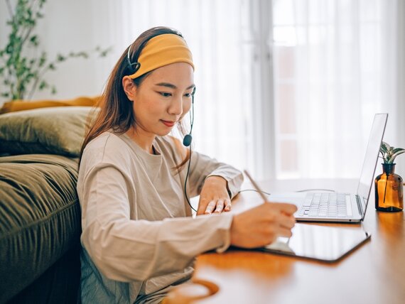 Eine Frau mit Headset sitzt vor ihrem Laptop und macht sich Notizen.