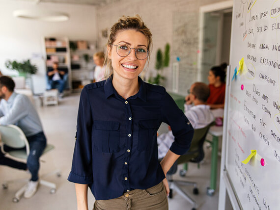 Eine junge, fröhliche Frau mit Brille steht vor einem Whiteboard in einem gemütlichen Grußraumbüro