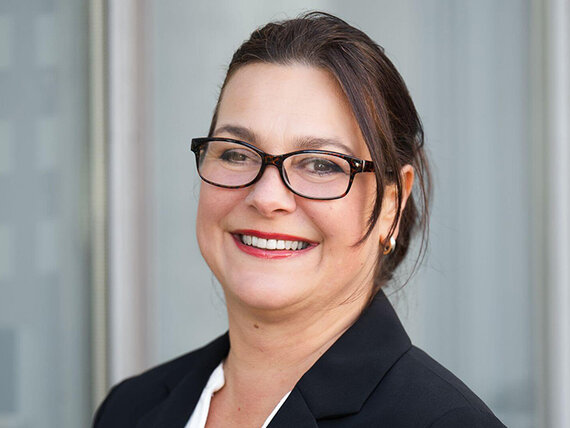 Eine lächelnde Frau mit Blazer, Brille und dunklen Haaren.
