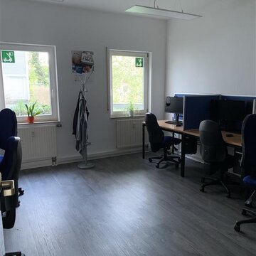 Raum mit 2 Fenstern und mehreren Arbeitsplätzen. Zwischen den Fenstern steht ein Kleiderständer. Rechts 2 Arbeitsplätze mit jeweils einem Bürostuhl, 2 Bildschirmen und Tastatur.