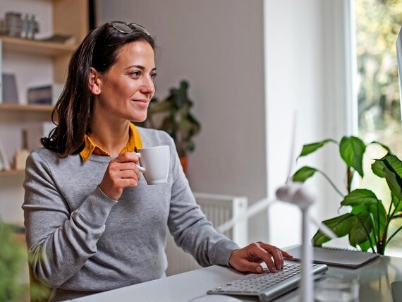 Eine Frau sitzt vor ihrem Computer und hält eine Kaffeetasse in der Hand.