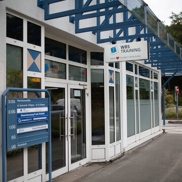 Außenansicht des Gebäudes. Glasfront mit blauem Überdachungsgestell. Schild mit der Aufschrift WBS TRAINING – Unser Herz schlägt für Bildung.