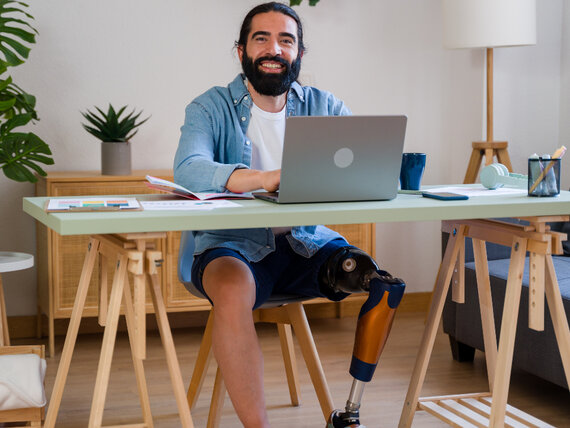 Mann mit Beinprothese sitzt an seinem Schreibtisch und arbeitet gut gelaunt am Laptop.