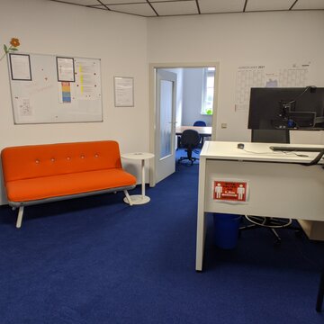 Büroraum mit einem weißen Schreibtisch auf dem 2 Bildschirme stehen, einem orangenen Sofa, einer magnetischen Pinnwand an der Wand sowie eine Druckstation und Türen zu zwei weiteren Räumen.