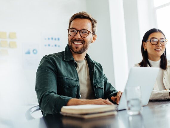 Eine smart aussehende Frau und ein junger fröhlicher Mann sitzen in einem Büroraum mit Whiteboard und Laptop