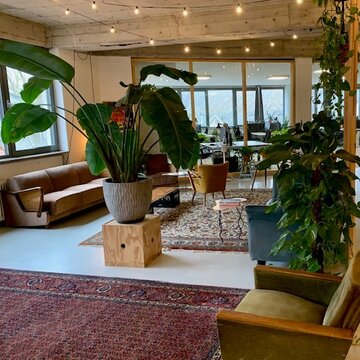 Gemütlicher Aufenthaltsraum mit Wohnzimmeratmosphäre: Sofa, Sesseln, Teppiche und große, grüne Zimmerpflanzen. An der Decke hängt eine Lichterkette.