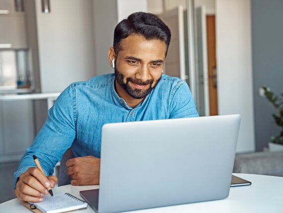 Ein Mann mit Kopfhörern in den Ohren sitzt vor seinem Laptop und macht sich Notizen.