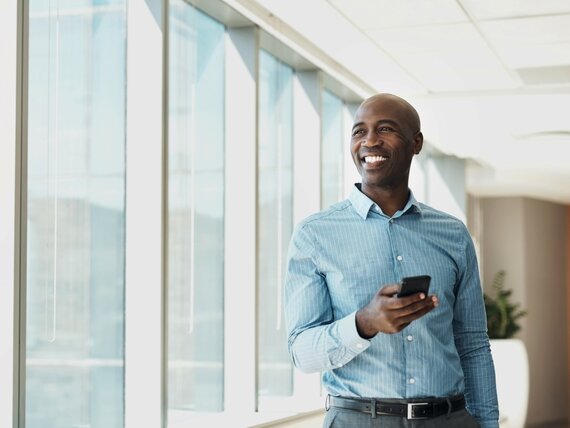 Ein fröhlicher Mann steht in einem Büro am Fenster mit seinem Smartphone in der Hand