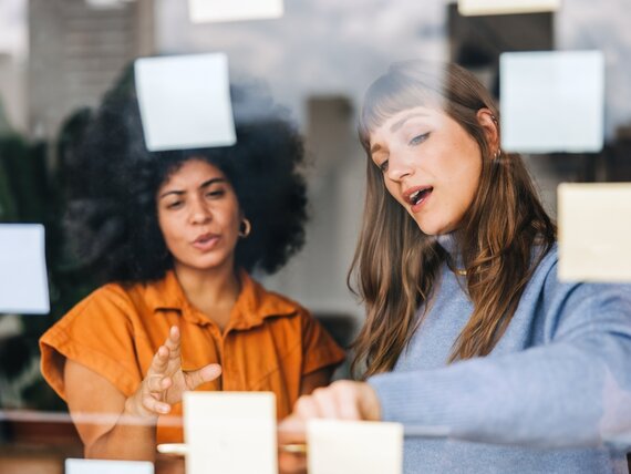 Zwei Frauen stehen vor einer Glaswand mit Notizzetteln und tauschen ihre Gedanken aus