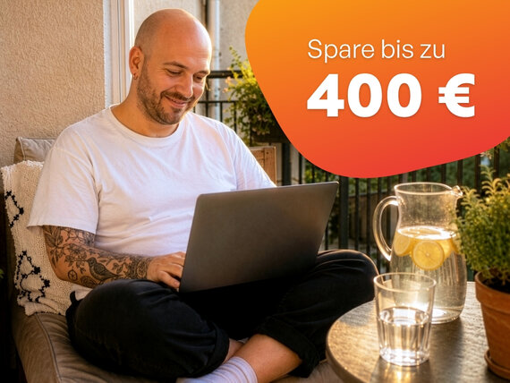 Person mit Tattoos sitzt auf Balkon und arbeitet mit Laptop auf dem Schoß neben Tisch mit Karaffe und Glas