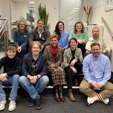 Teambild. 10 Personen sitzen im Flur auf einer Treppe und lachen fröhlich in die Kamera.