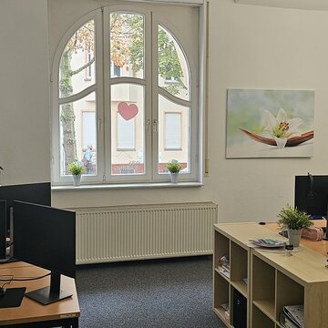 Schulungsraum mit mehreren Arbeitsplätzen mit jeweils 2 Bildschirmen, einem Bürostuhl, Tastatur, Maus und Headset. An den Wänden hängen Plakate und Bilder.