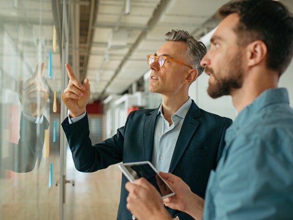 Zwei Männer besprechen Notizzettel, die an einer Glaswand kleben.